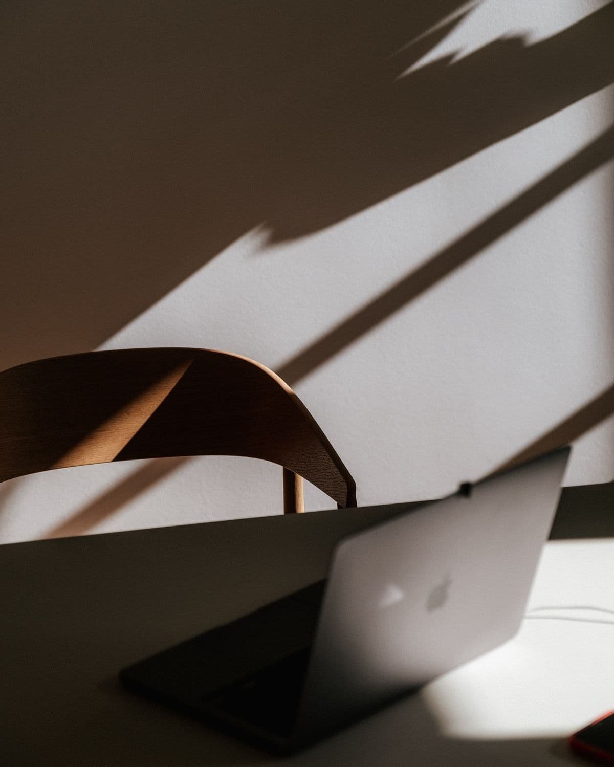 Laptop on a desk with a wooden chair, featuring dramatic sunlight and sharp shadows.