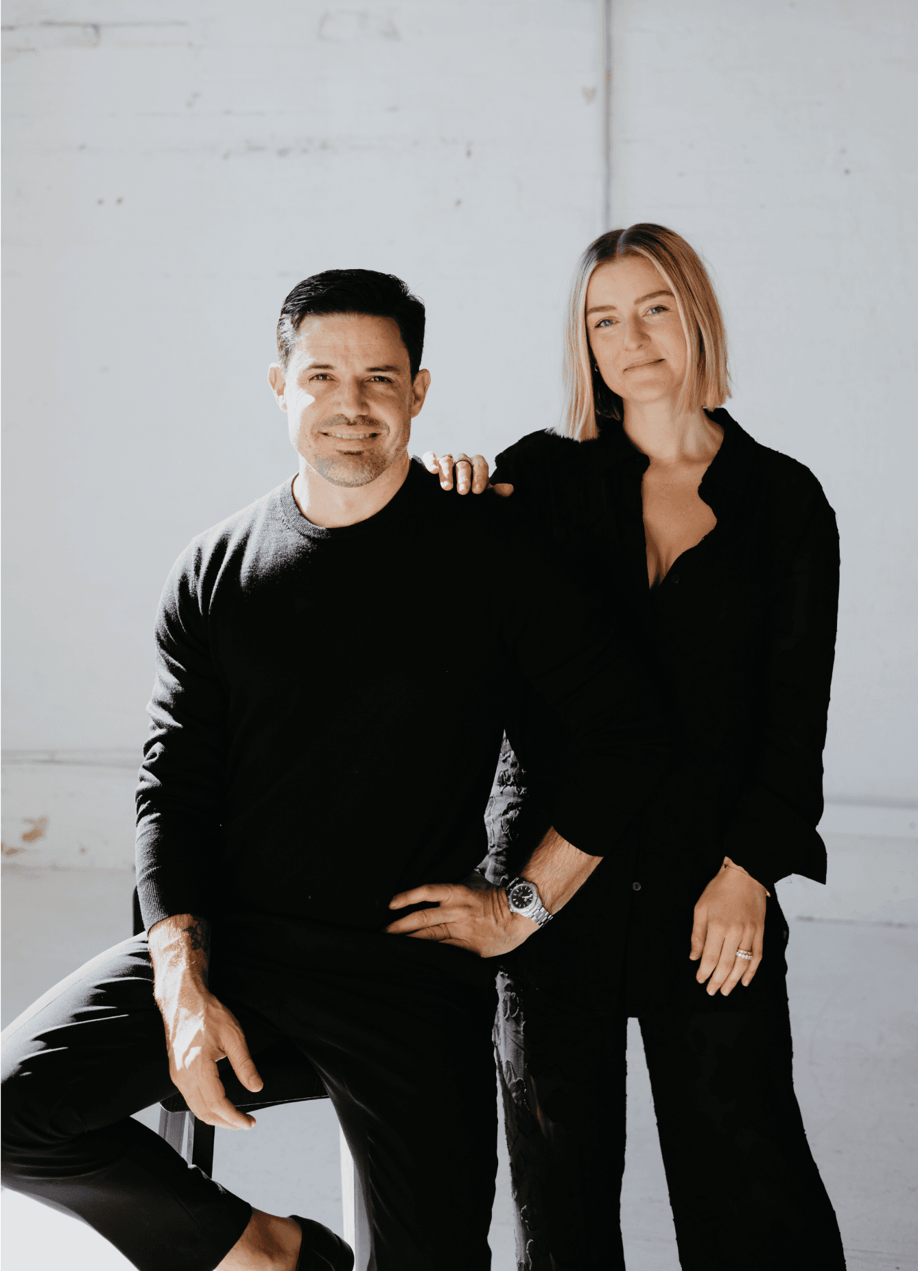 A man and woman in black outfits pose together against a minimalist white background.